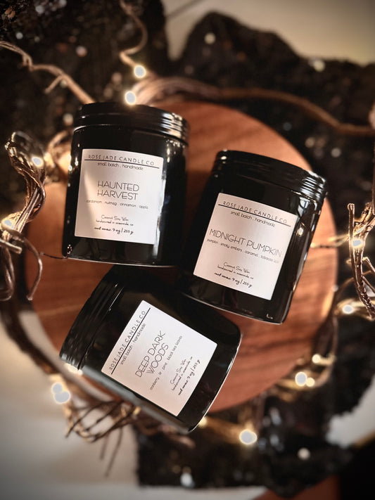 Three black ceramic container candles with black lids and white square  labels on a wooden tray against a blurred background