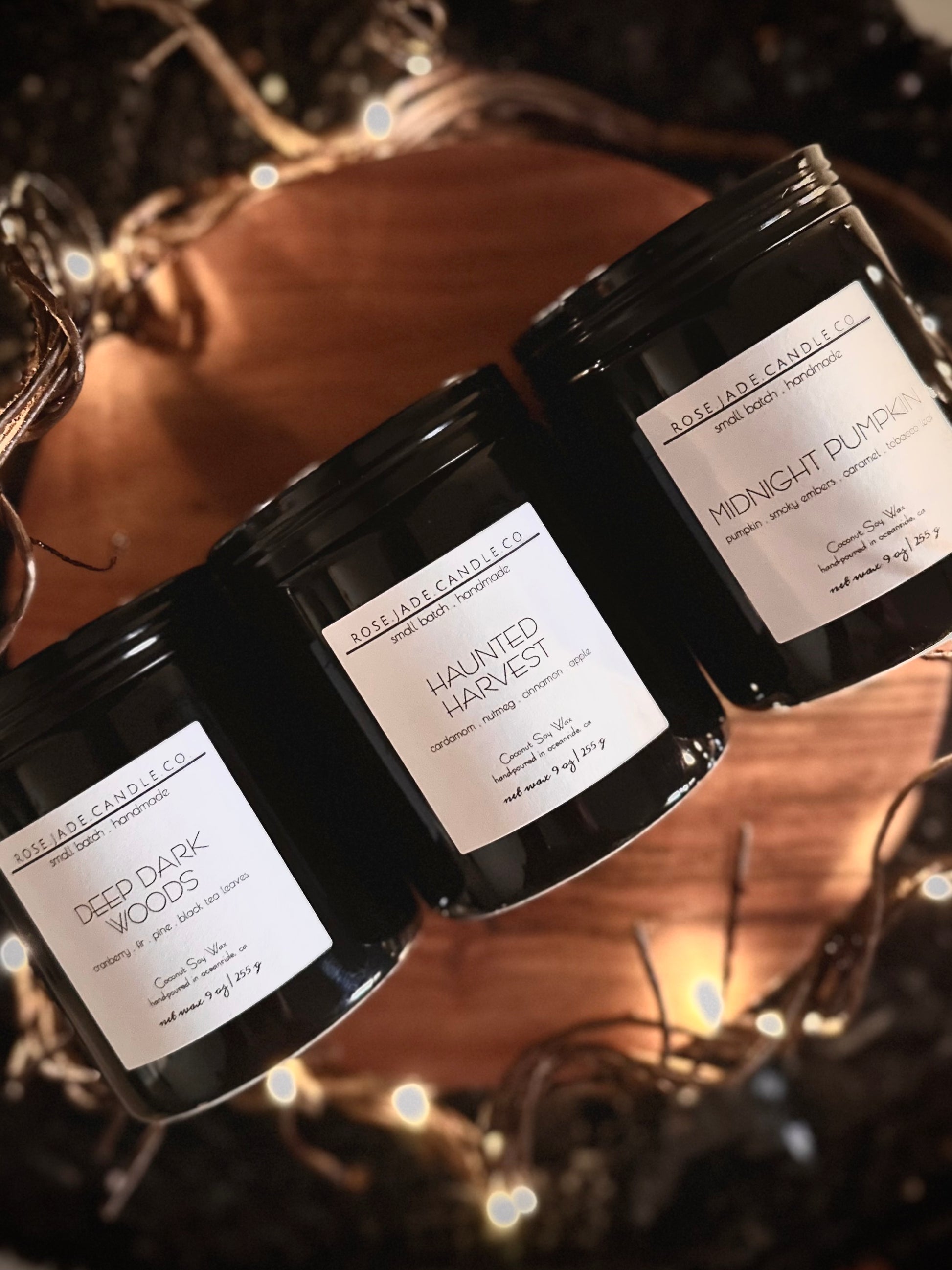 Three black ceramic container candles with black lids and white square  labels on a wooden tray against a blurred background
