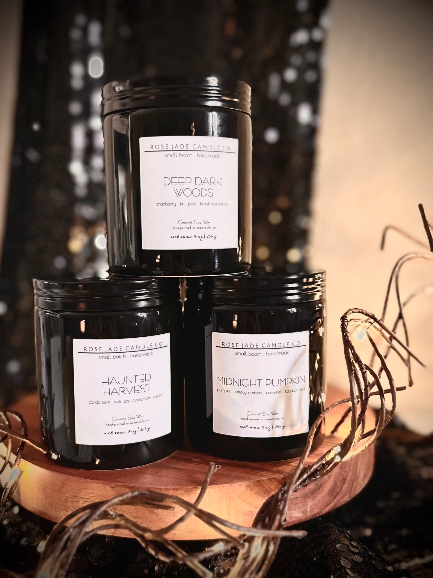 Three black ceramic container candles with black lids and white square  labels on a wooden tray against a blurred background