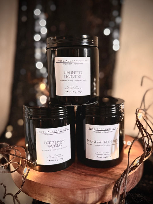 Three black ceramic container candles with black lids and white square  labels on a wooden tray against a blurred background